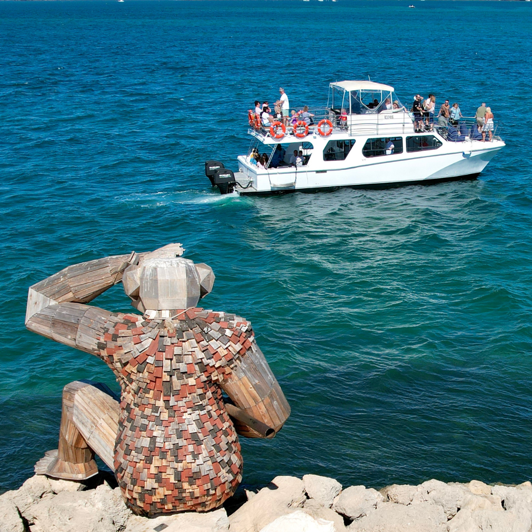 one of Thomas Dambo's wooden Giants of Mandurah sculptures sitting on a rock, watching out of the water, with a boat in the background driving past