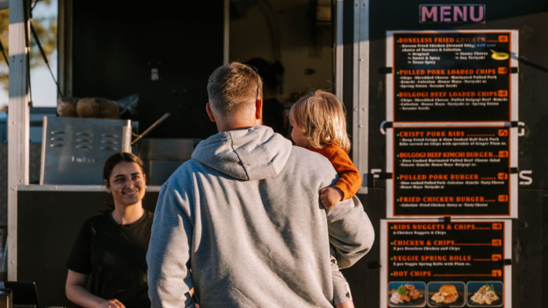 A father with his little child on his arm is standing in front of a food truck talking to a young lady who is taking his order