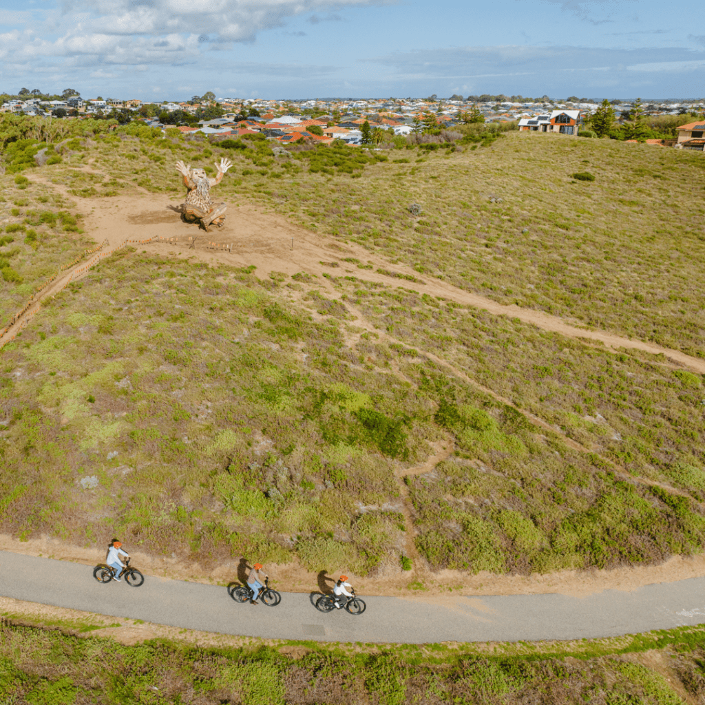 3 cyclists on e-bikes riding on a shared path with one of Thomas Dambo's Giants of Mandurah in the background