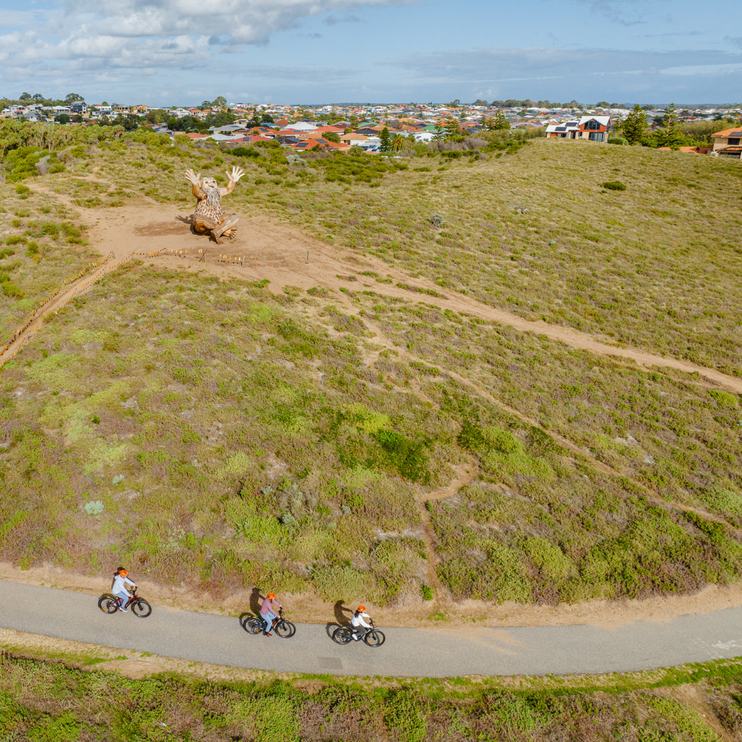 3 cyclists on e-bikes riding on a shared path with one of Thomas Dambo's Giants of Mandurah in the background