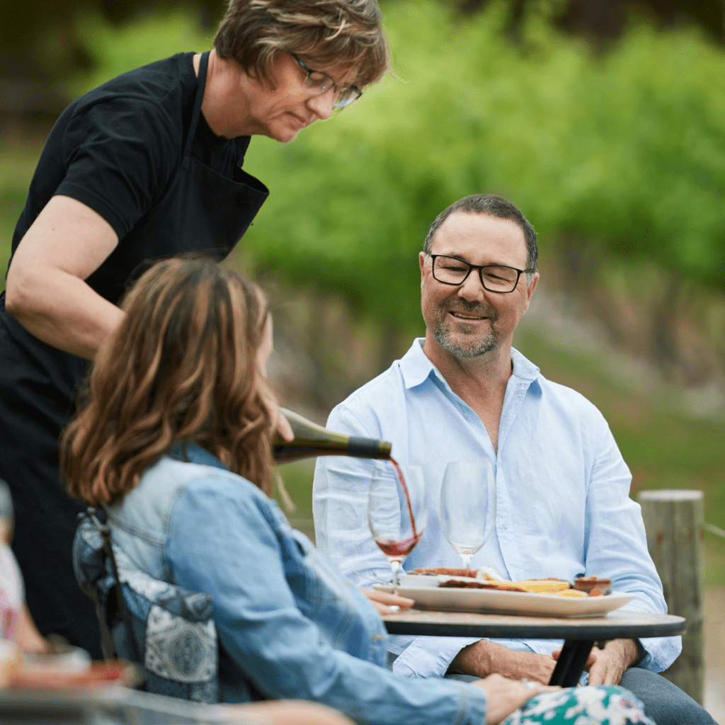 a woman serving wine to a couple at an outdoor table in a vineyard