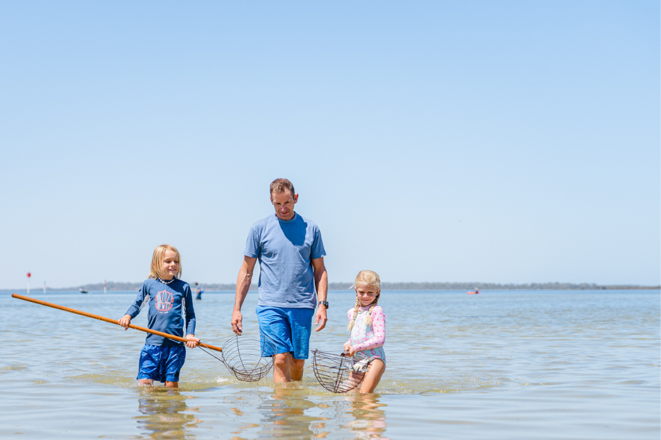 Two children and their Dad are walking in the water carrying crab scoops