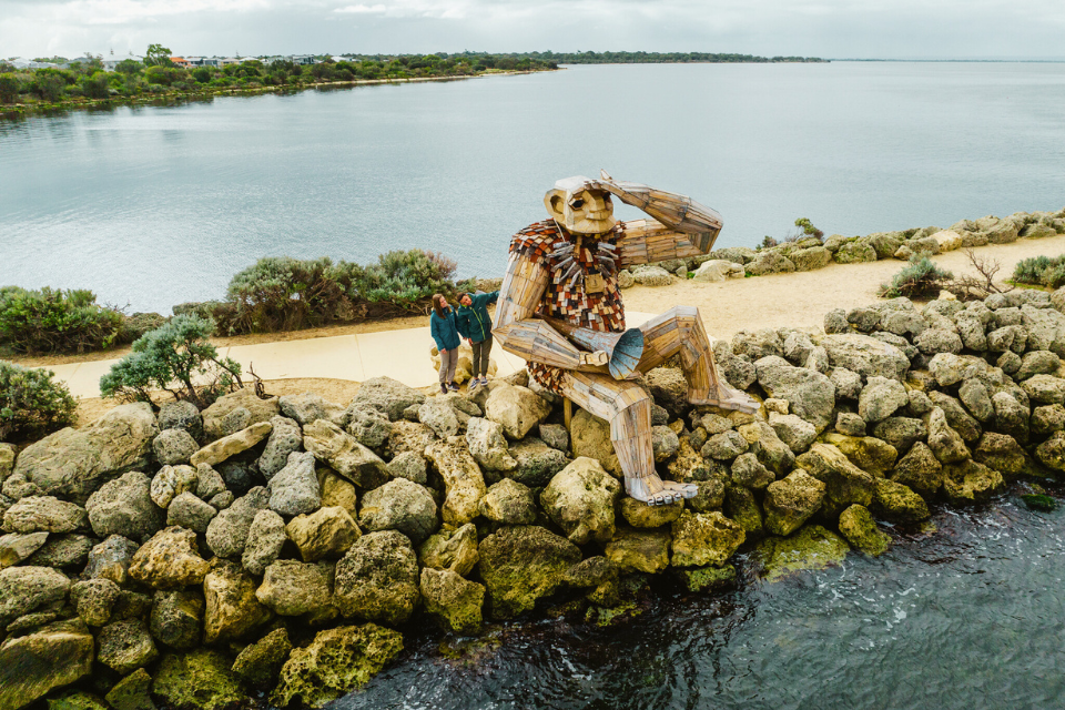 a wooden statue of a Troll sitting on a rock by the water